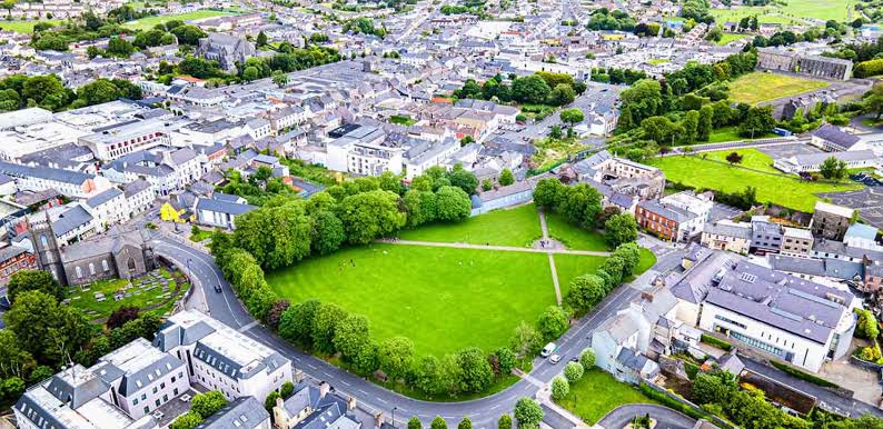 Aerial view of a circular green park with trees around it, intersected by crisscrossing walking paths, surrounded by buildings and roads in a small town.