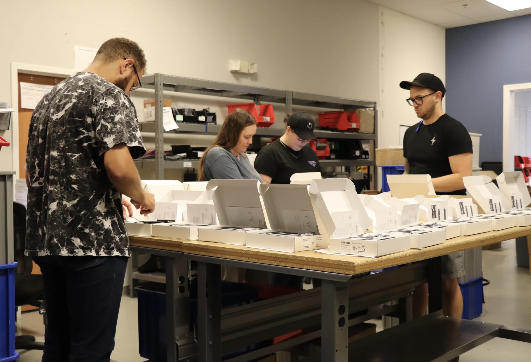 Four people working at a table with white boxes in an industrial workshop setting
