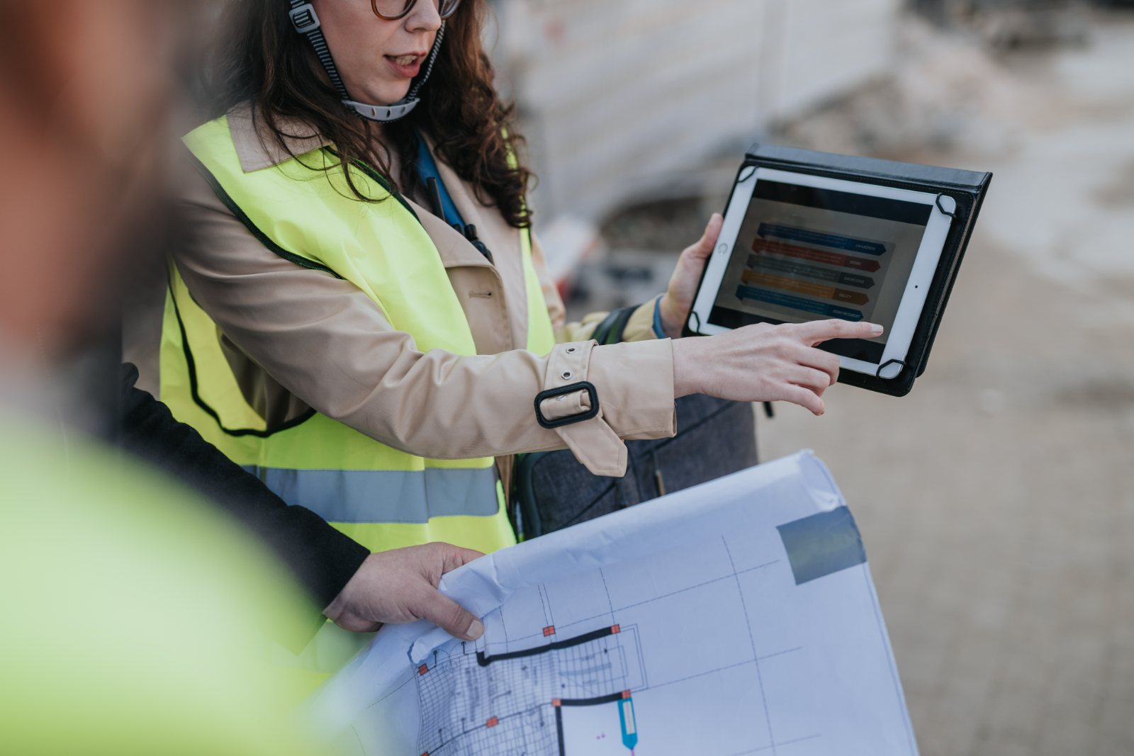 Female engineer in safety vest reviewing construction blueprint and digital tablet on site