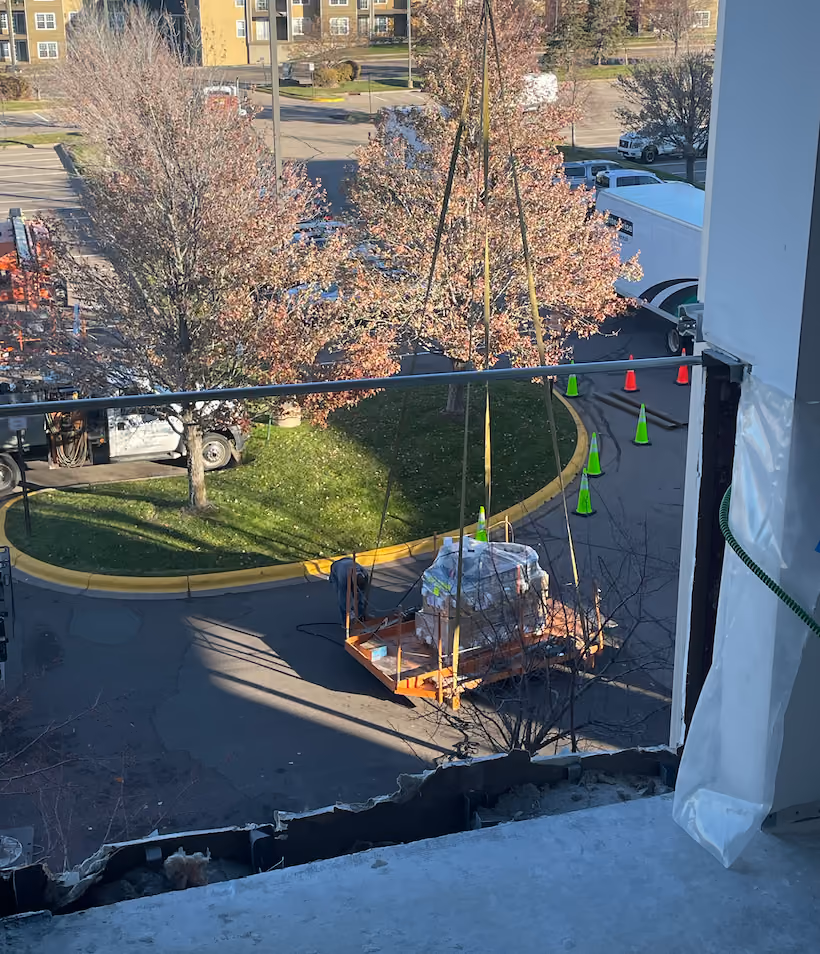 Construction crane lifting equipment wrapped in plastic outside near trees and parked vehicles.