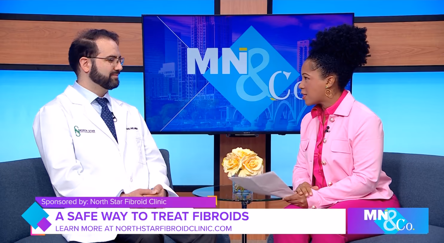 Doctor in white coat and host in pink jacket seated, discussing fibroid treatment on studio show.