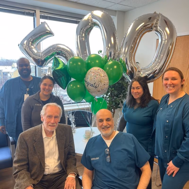 Six people in medical scrubs and a man in a suit celebrate with 500 silver balloons and green balloons.