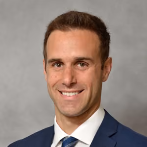 Smiling man with short brown hair wearing a blue suit, white shirt, and striped tie.