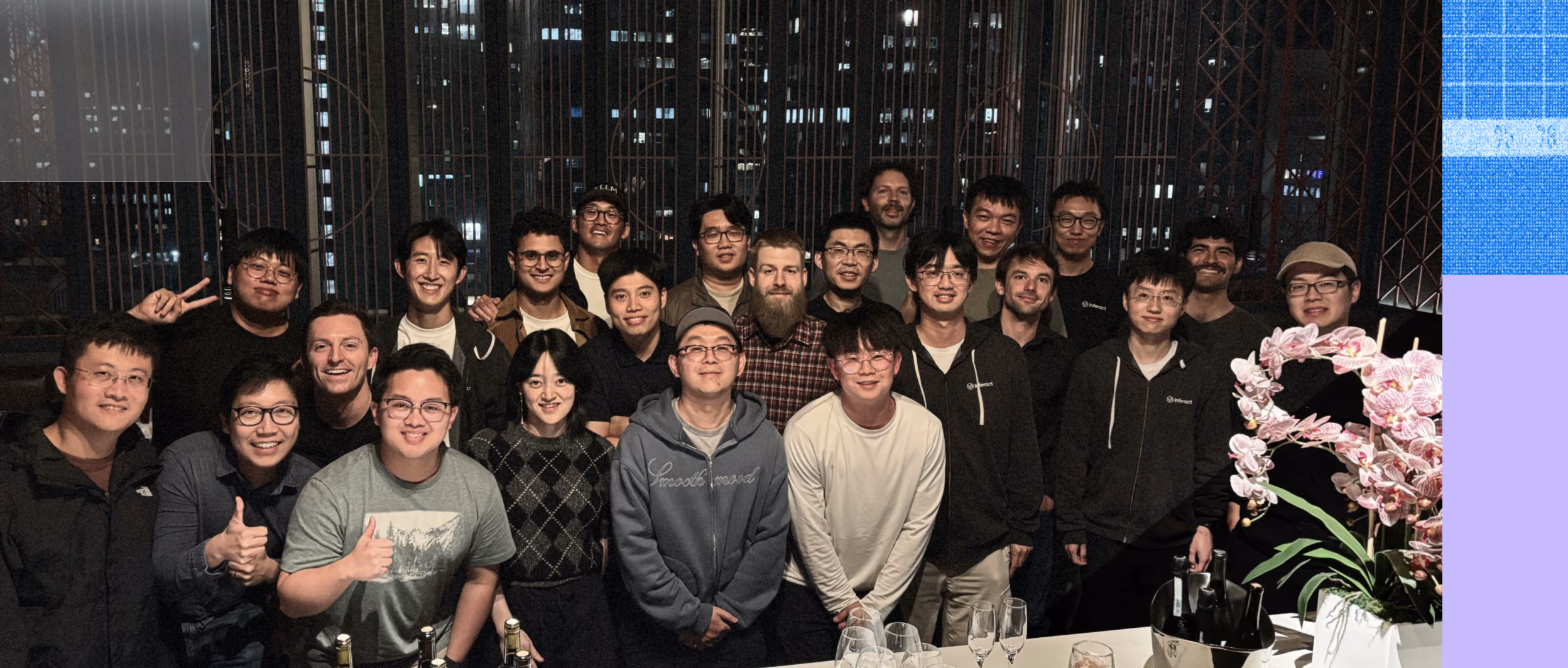 Group photo of smiling Inferact employees posing indoors at night in front of large windows showing city lights, with a table in front holding flowers, wine glasses, and bottles.