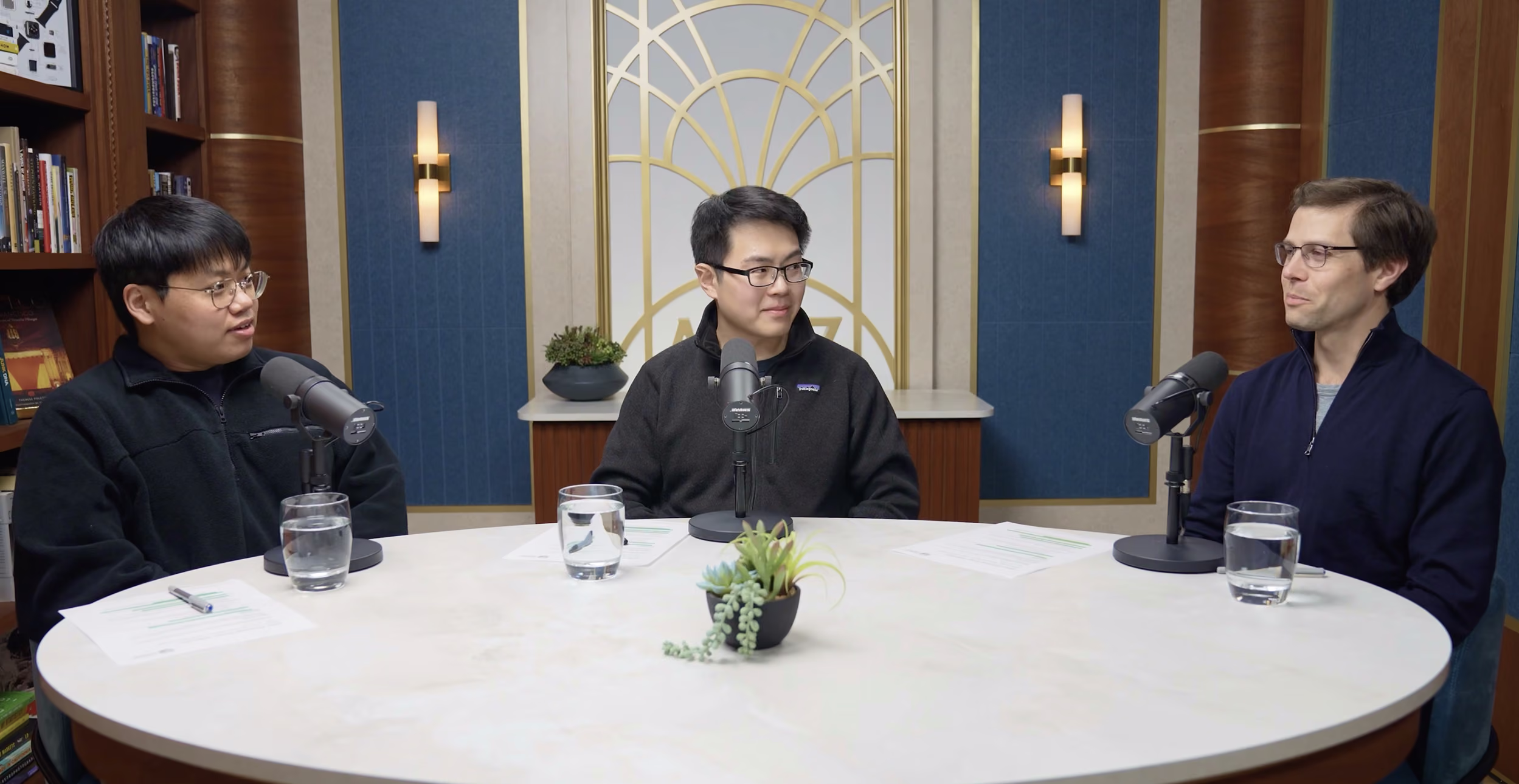 Three men seated around a round table engaged in conversation with microphones in front of them, glasses of water on the table, and a decorative plant centerpiece, in a room with blue and brown walls and wall sconces.