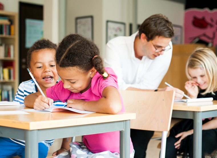 Young students writing in notebooks while a teacher helps