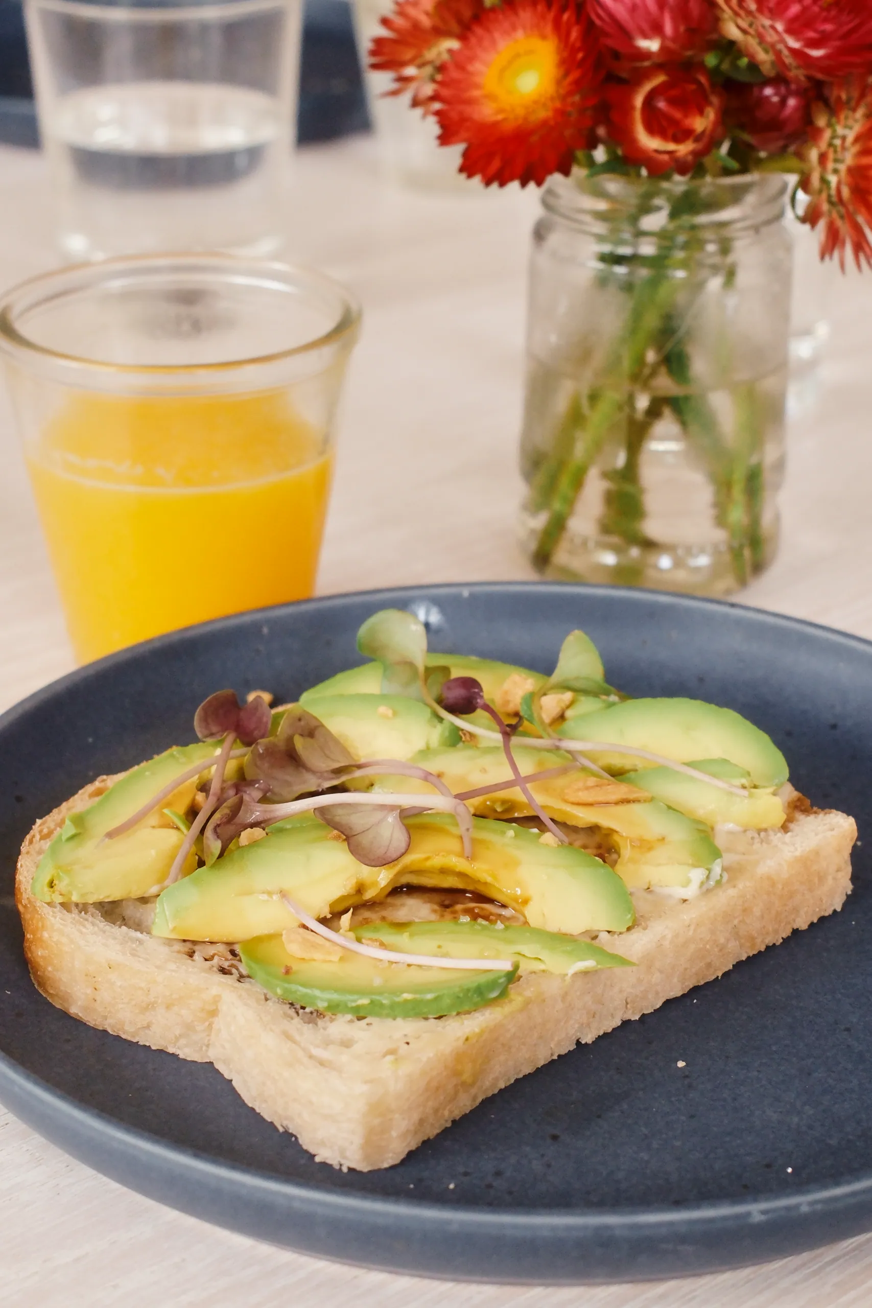 Avocado toast topped with microgreens on a blue plate next to orange juice and strawflowers