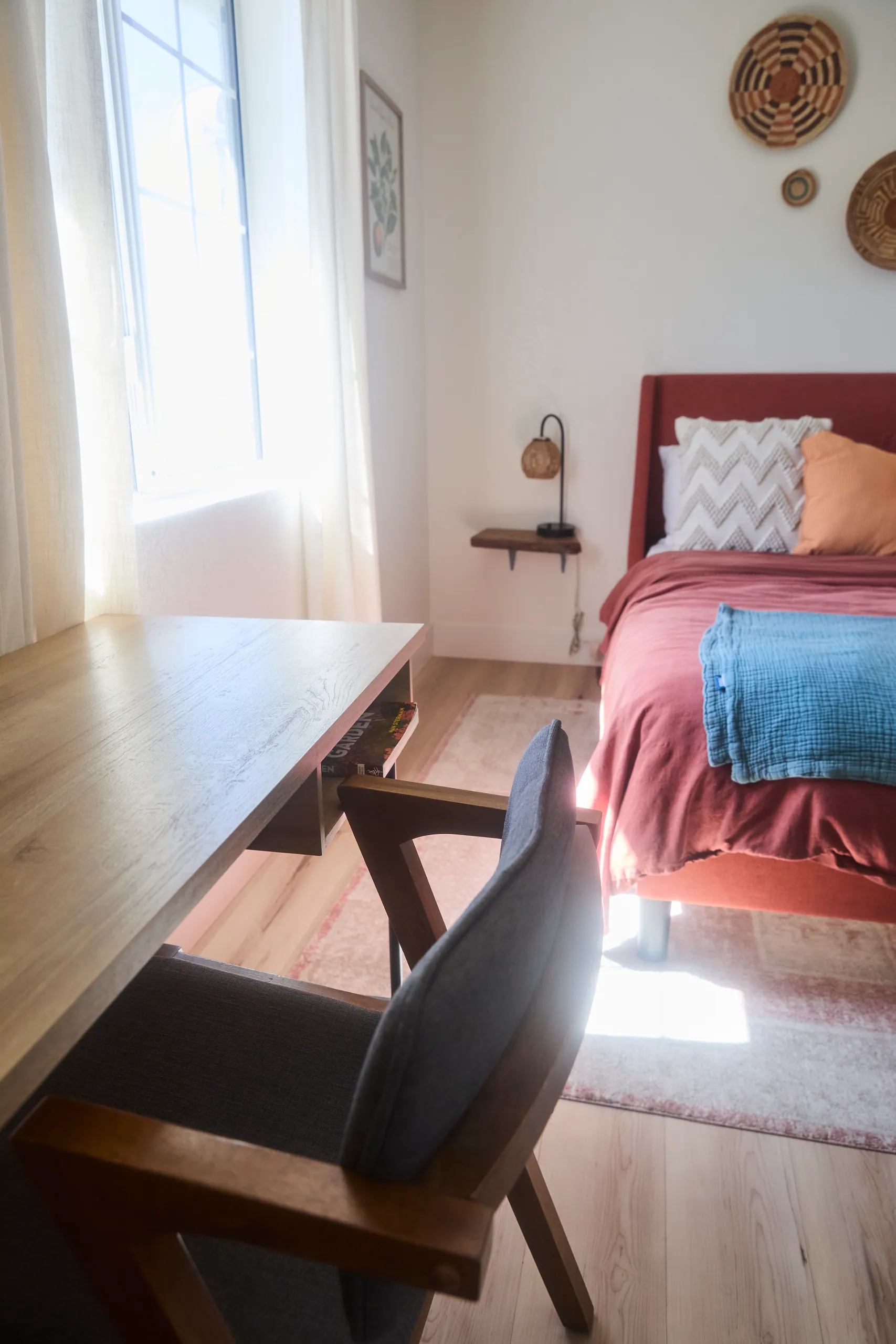 Wooden writing desk and chair in a sunlit guest bedroom beside a rust-red bed