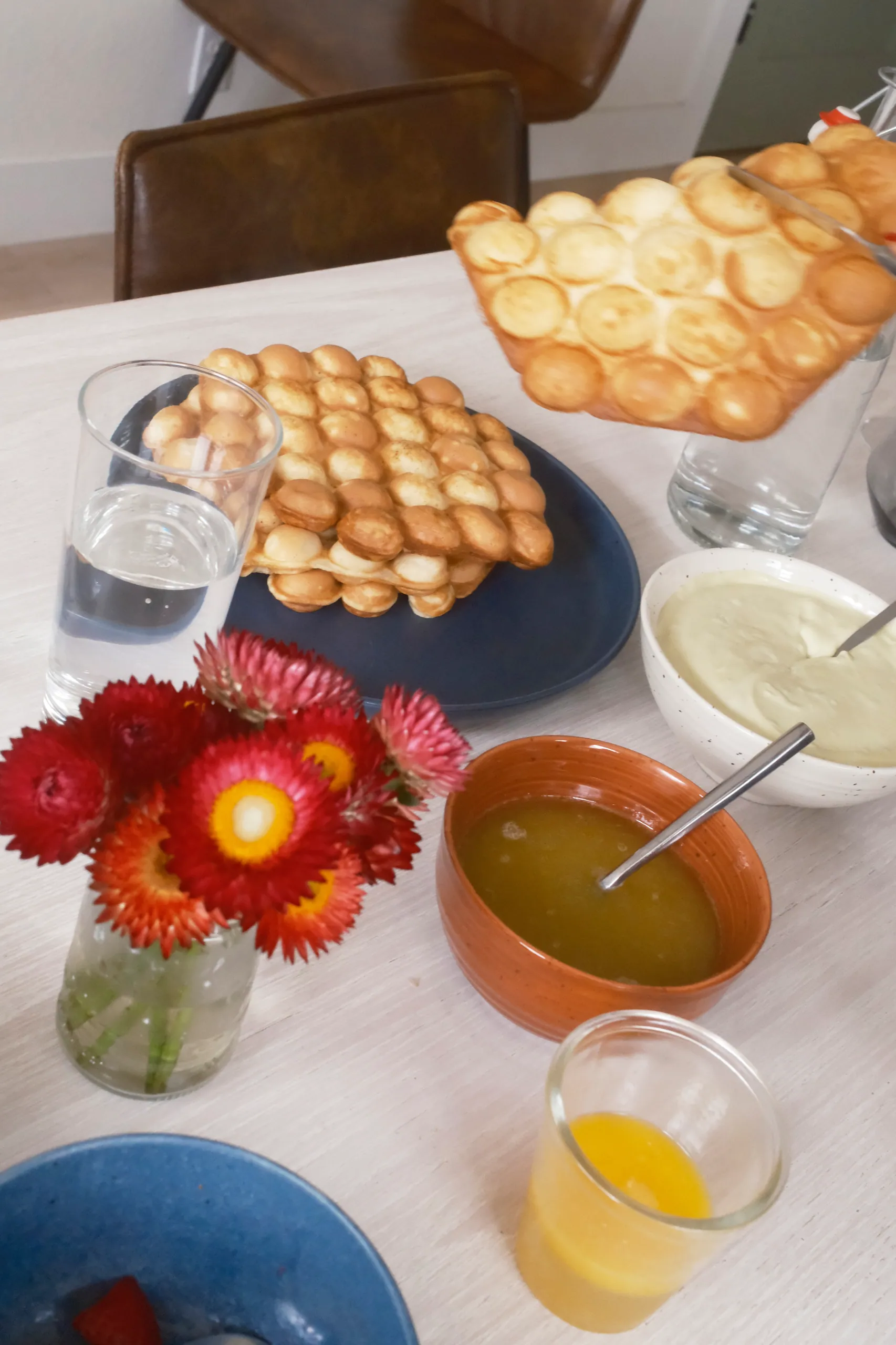 Golden bubble waffles on a blue plate with dipping sauces, orange juice and red strawflowers