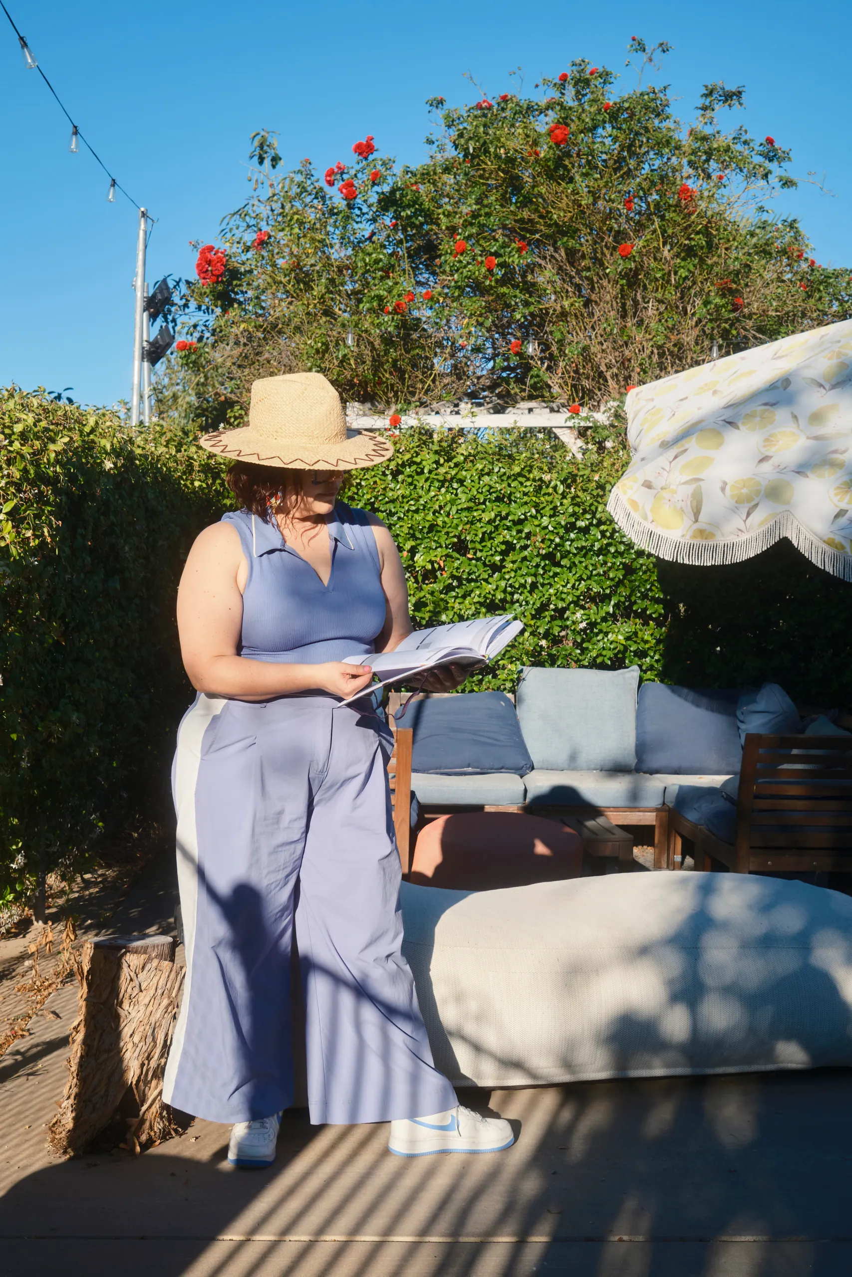 Woman in a straw hat and lavender jumpsuit reading on a sunny patio with a lemon-print umbrella