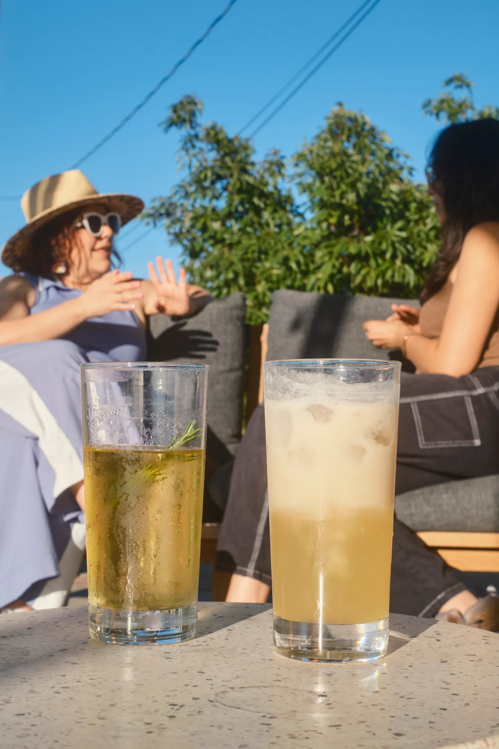 Two iced drinks on a terrazzo table with guests chatting on the patio behind them