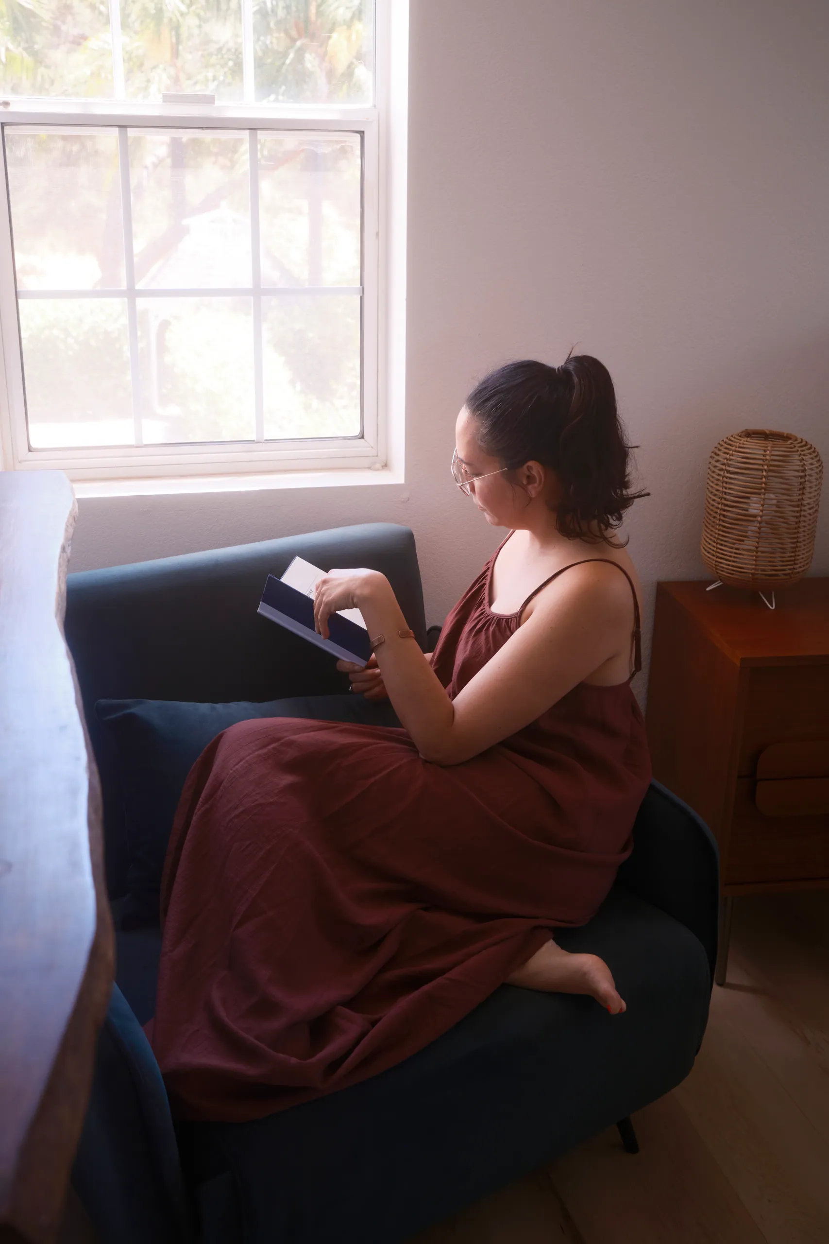 Woman reading a book on a teal sofa beside a sunny window and a rattan lamp