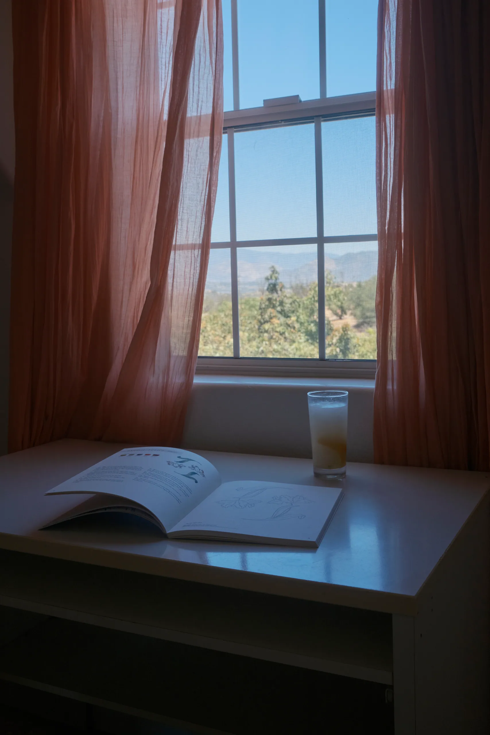 Sunlit writing desk with an open sketchbook and iced drink beside a window overlooking the hills