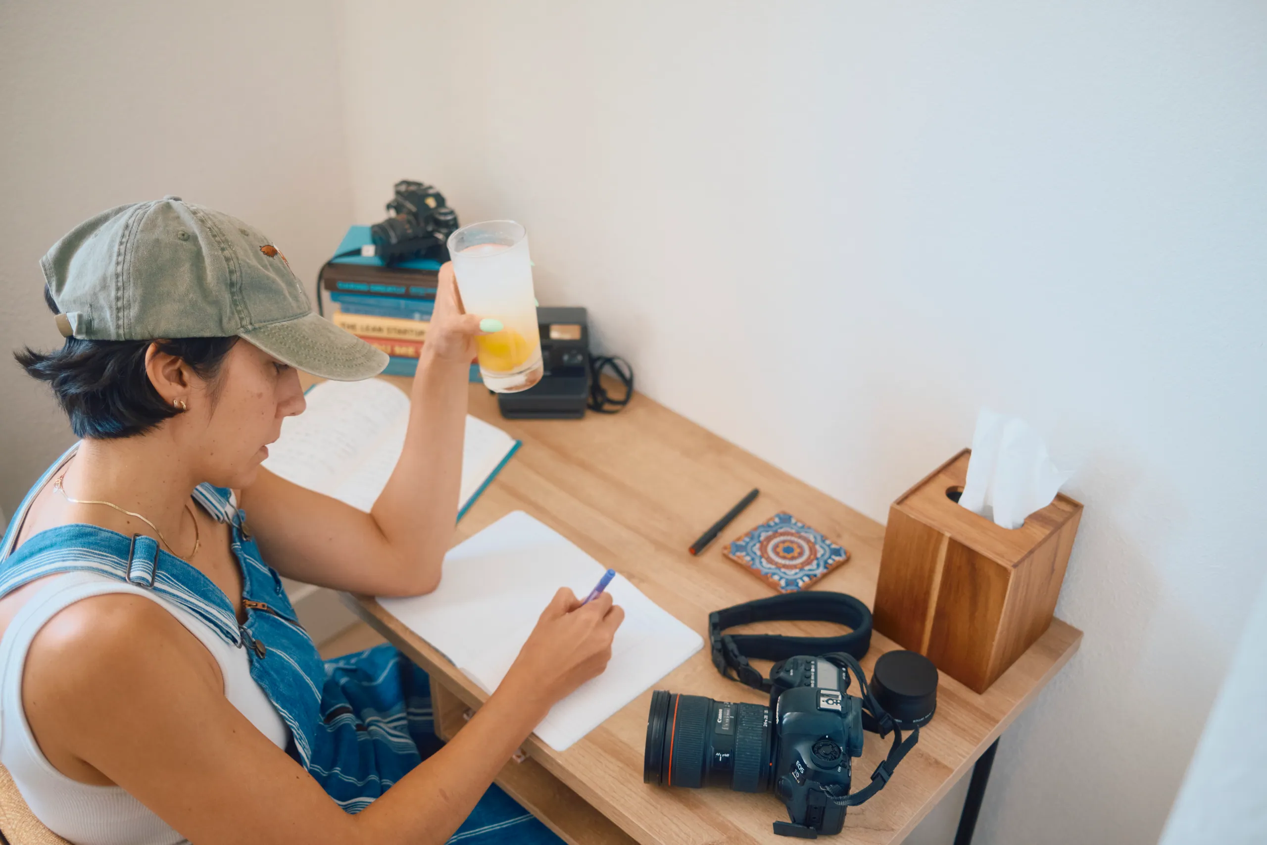 Woman in a cap and striped overalls writing in a notebook at a wooden desk beside a camera