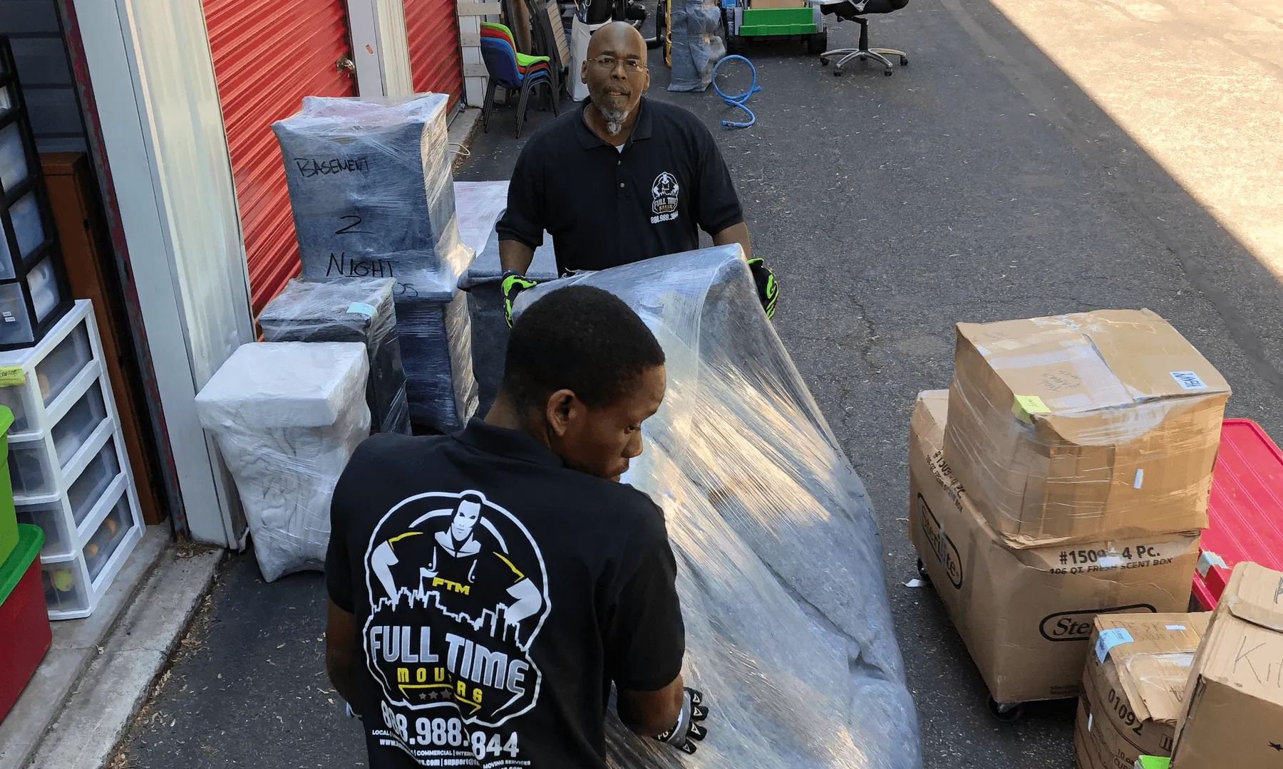 Two Full Time Movers crew members moving plastic-wrapped items past stacked boxes