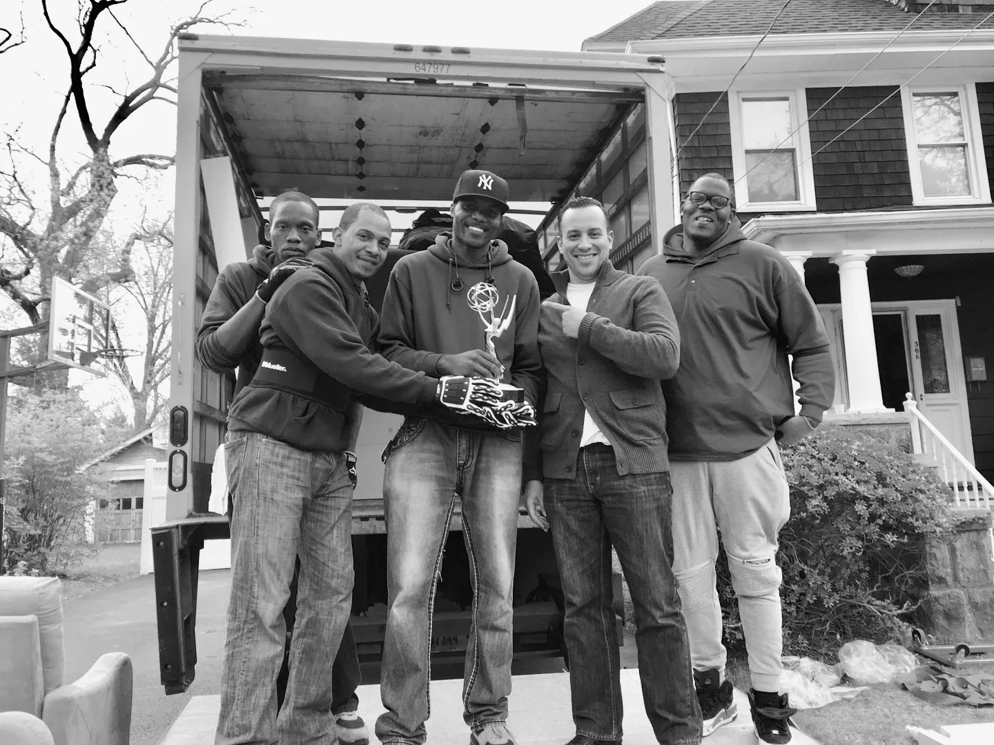 Black-and-white photo of five men posing in front of an open moving truck, one holding a trophy