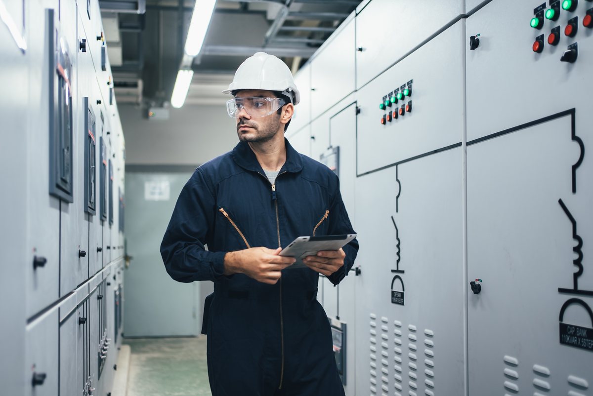 Engineer in a hard hat holding a tablet beside electrical cabinets