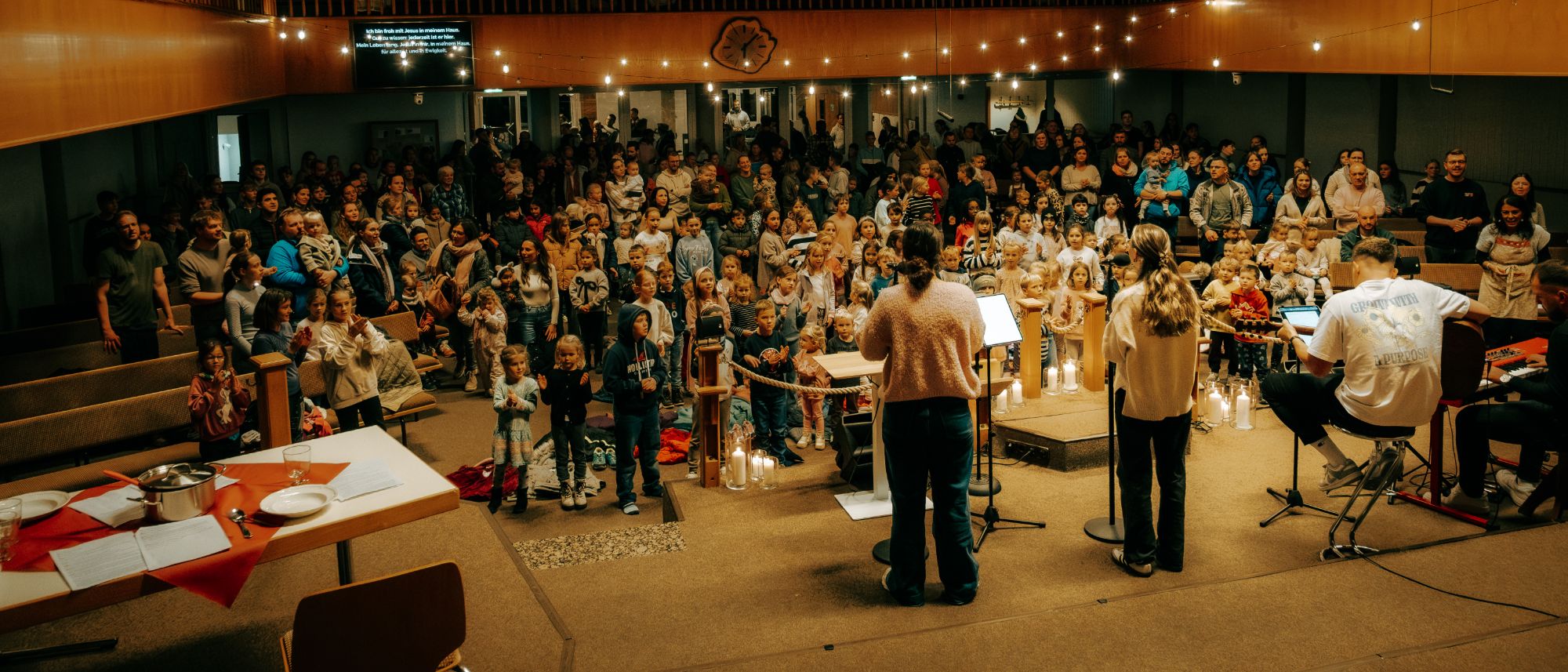Eine Band mit Sängerinnen, Gitarre und Keyboard spielt beim Lichterfest, Kinder und Familien im vollen Saal singen und klatschen mit