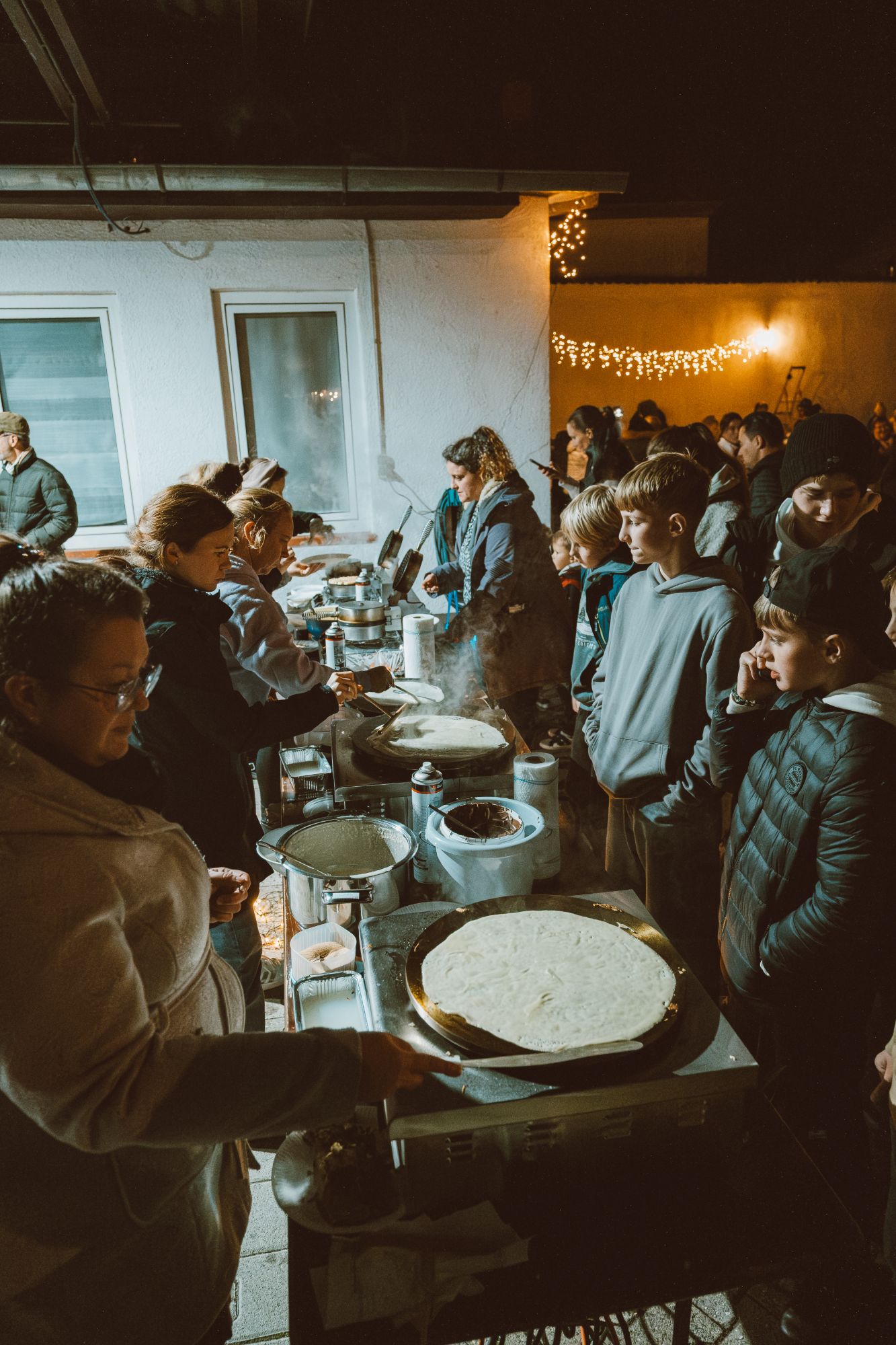 Helferinnen backen beim Lichterfest frische Crêpes, Kinder und Jugendliche warten unter Lichterketten auf ihr Essen