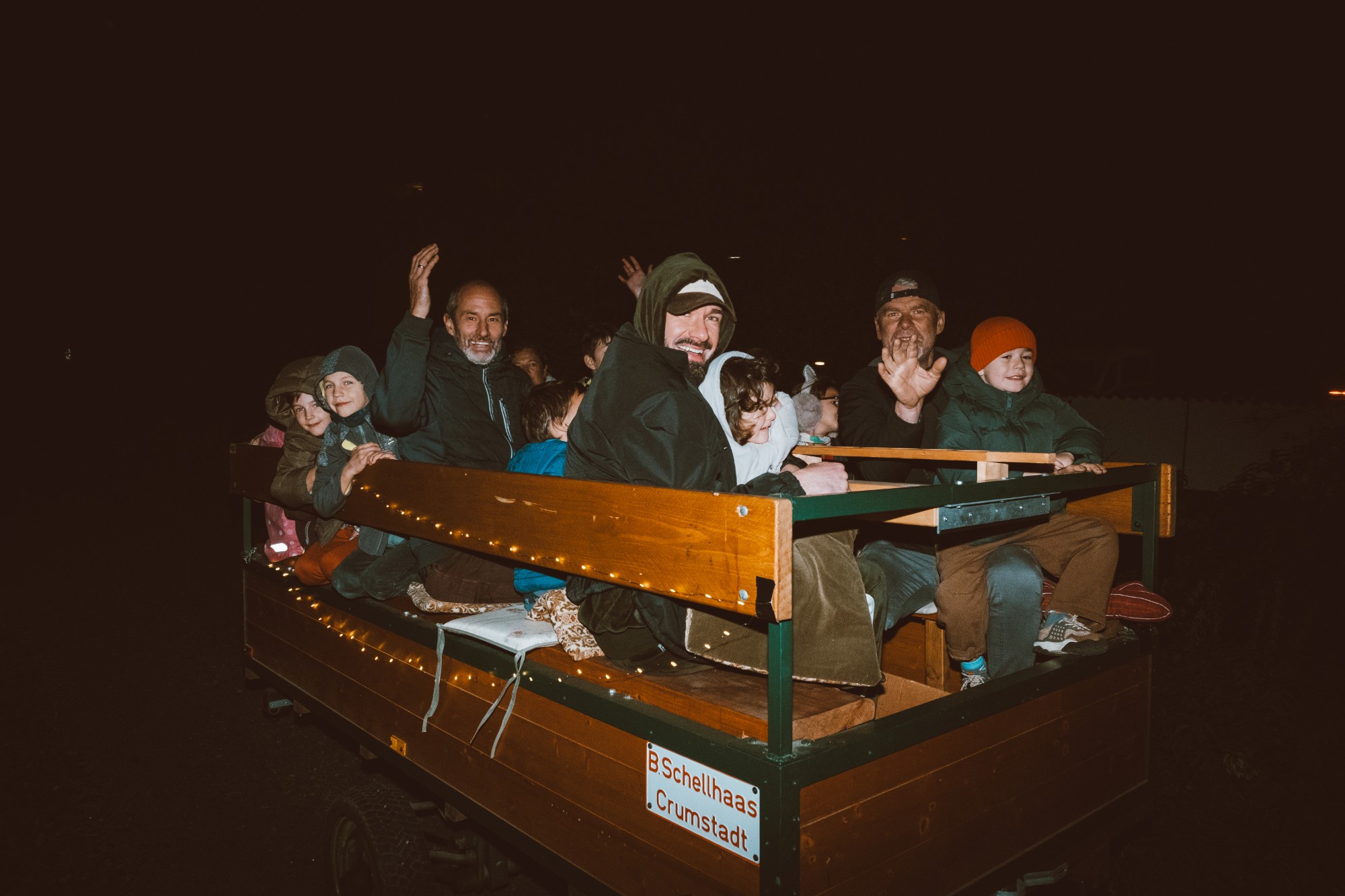 Kinder und Erwachsene winken lachend vom beleuchteten Anhänger bei der abendlichen Traktorfahrt des Lichterfests