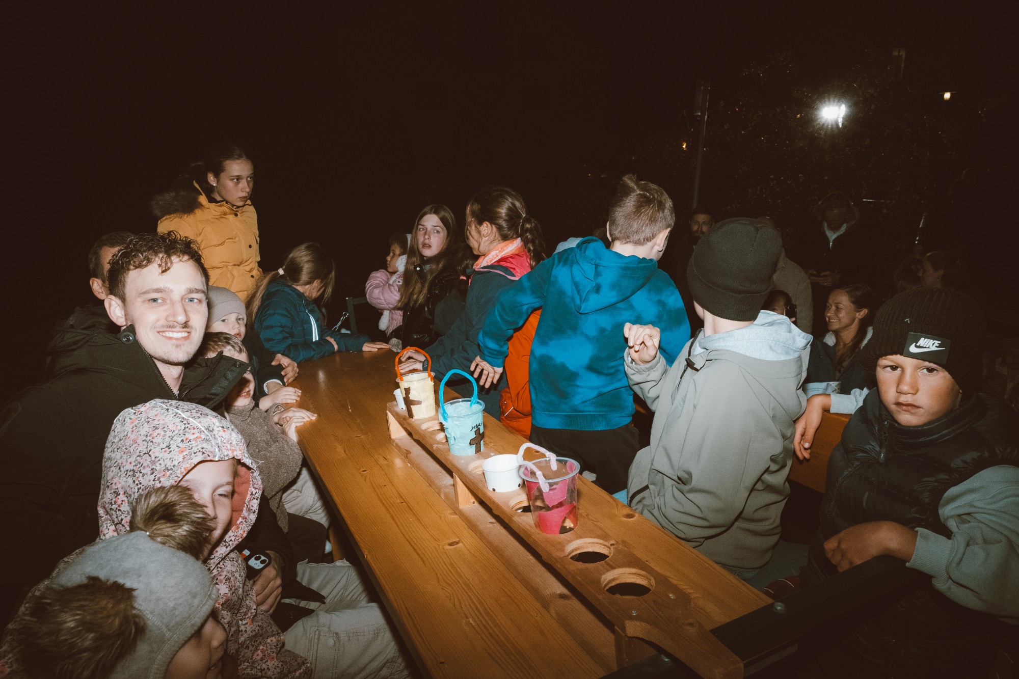 Selbstgebastelte Windlichter leuchten auf einem Holztisch, drumherum Kinder und Familien