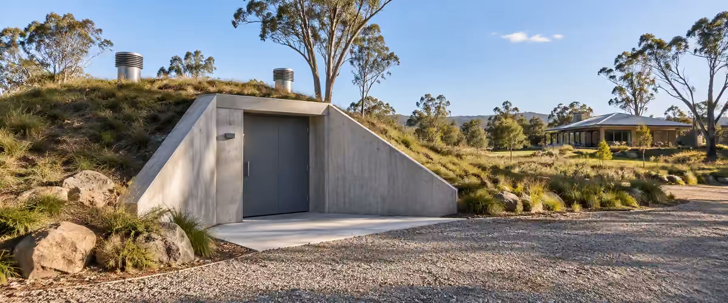 Earth-covered concrete bushfire shelter with a steel door, set into a grassy mound near a rural home