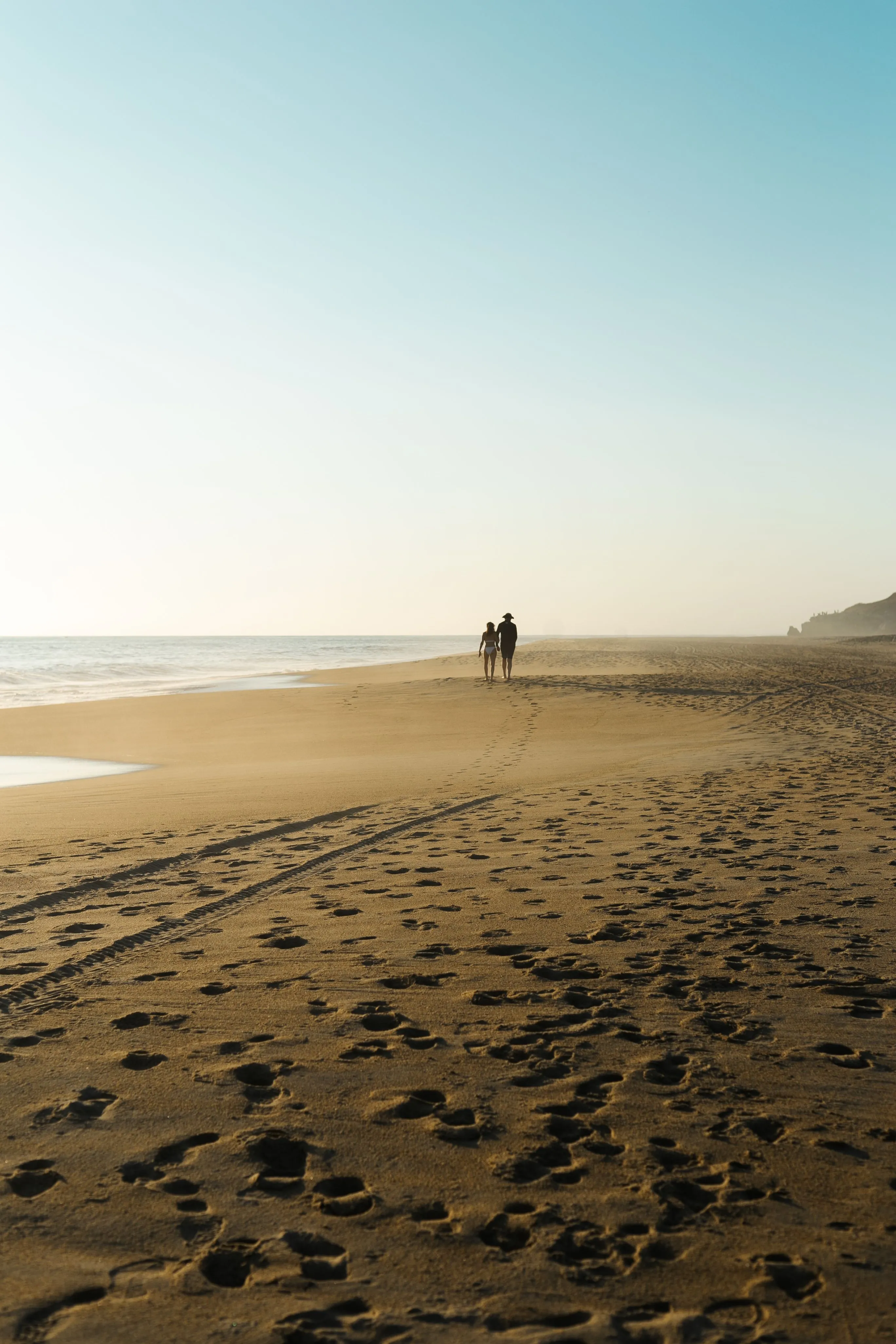 Couple walking on wide empty sandy beach with footprints at sunrise