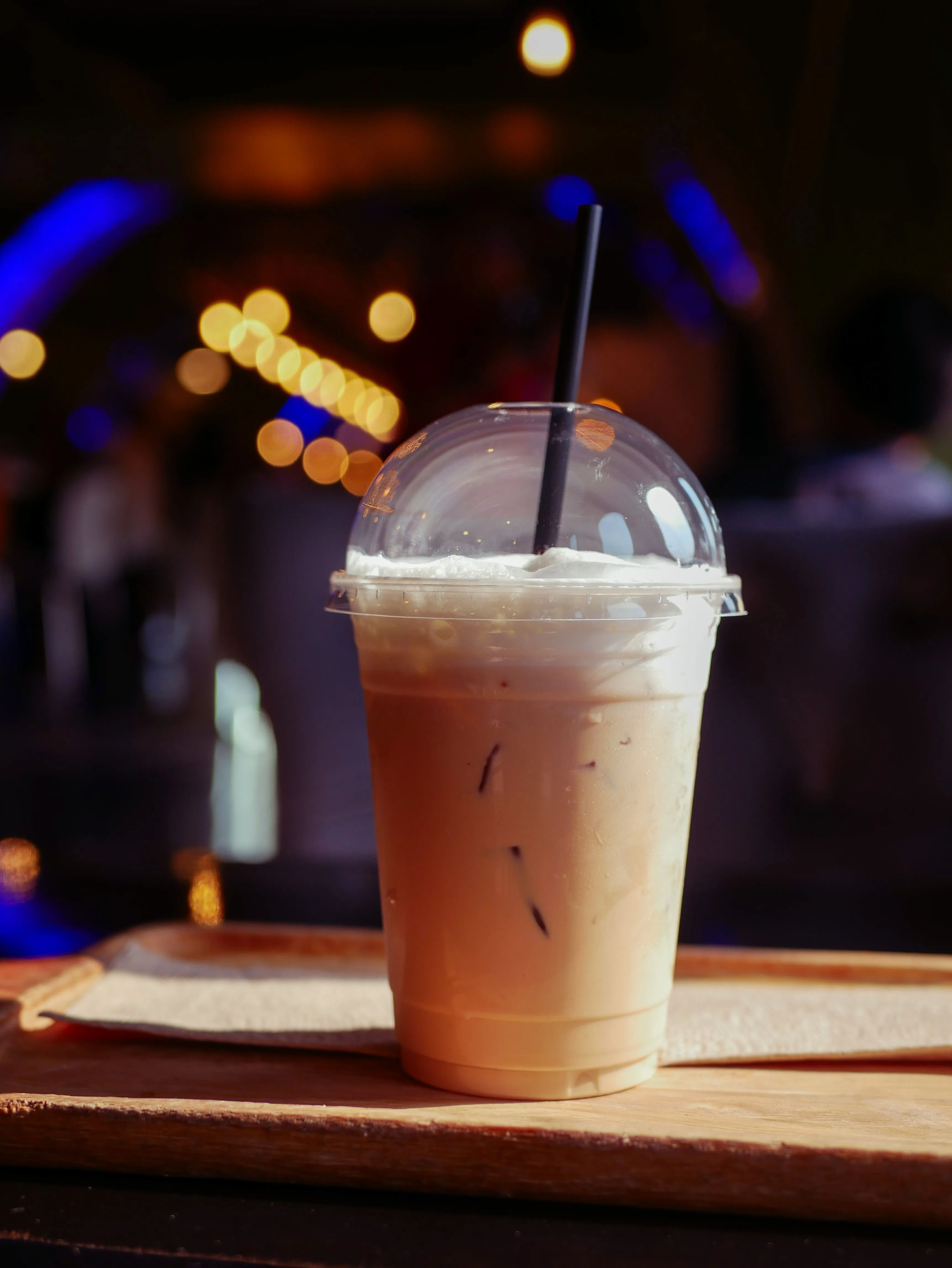 Iced latte in plastic dome-lid cup on wooden tray, bokeh lights behind