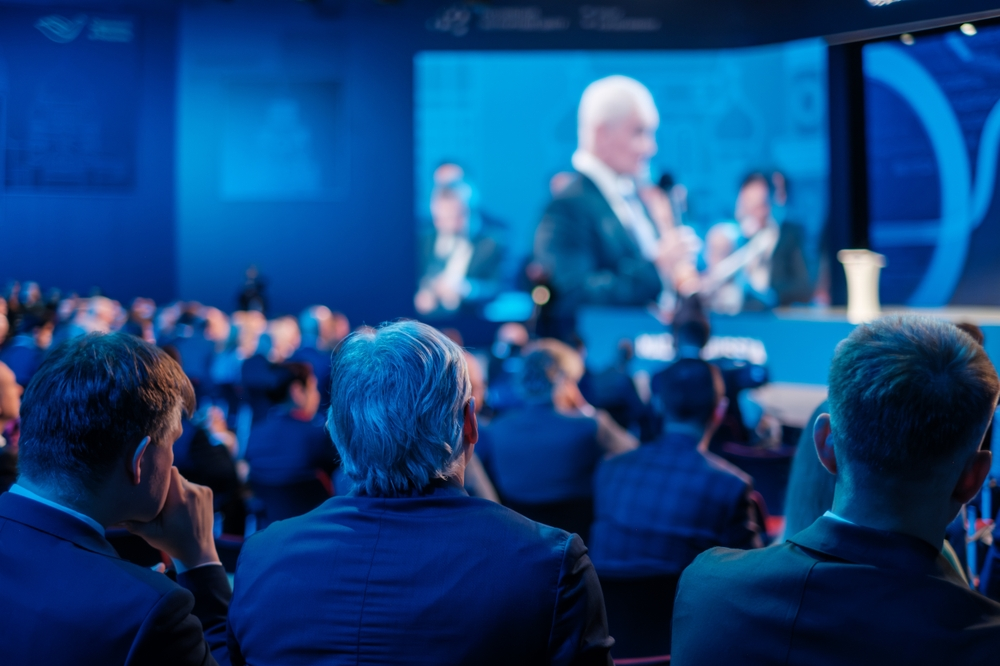 Blurred audience watches a speaker on stage, bathed in cool blue conference lighting.