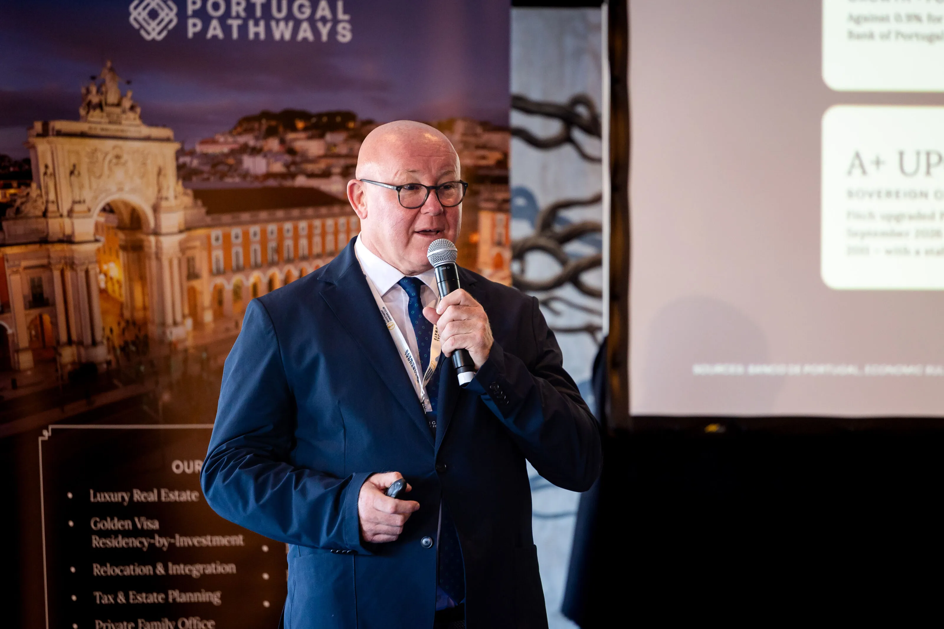 Man in glasses speaks into a microphone, wearing a navy suit and lanyard