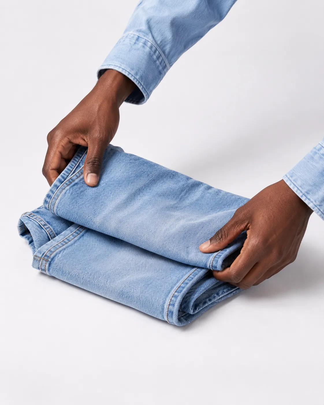 Hands folding a pair of washed jeans in a white studio.