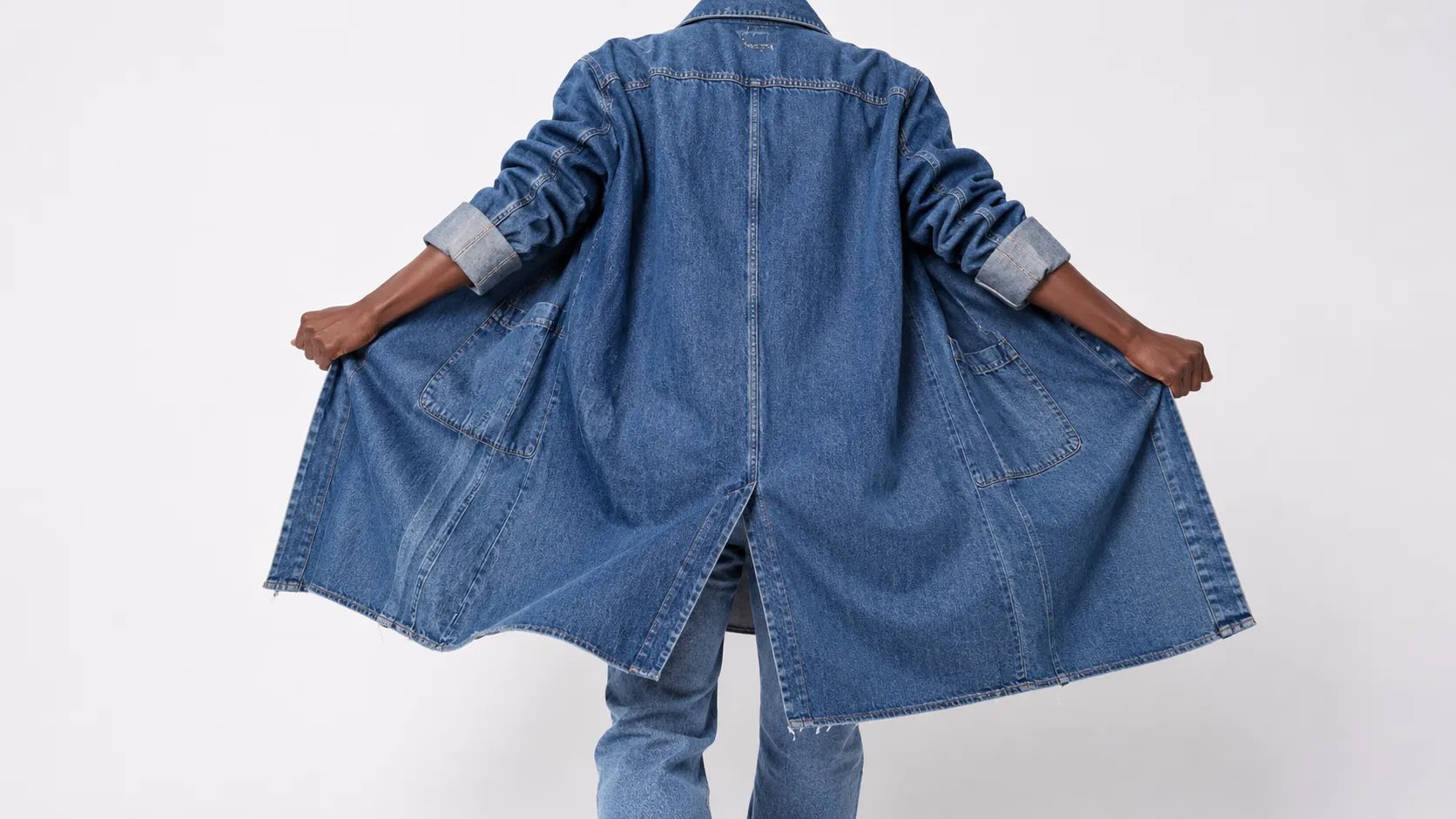 Rear view of a person holding open a long stone-washed denim coat in a white studio.