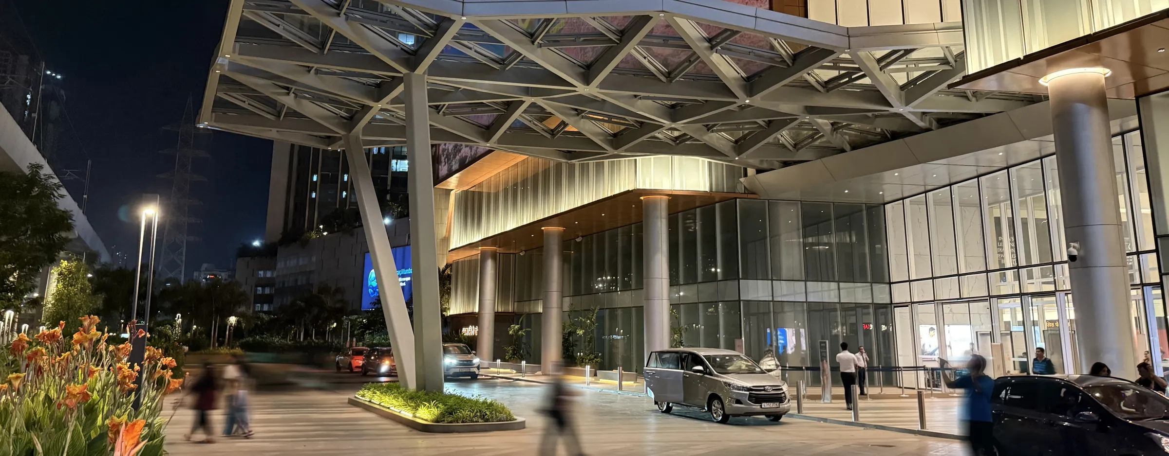 SkyCity Mall entrance at night, with its steel canopy, glazed facade and drop-off road, Mumbai