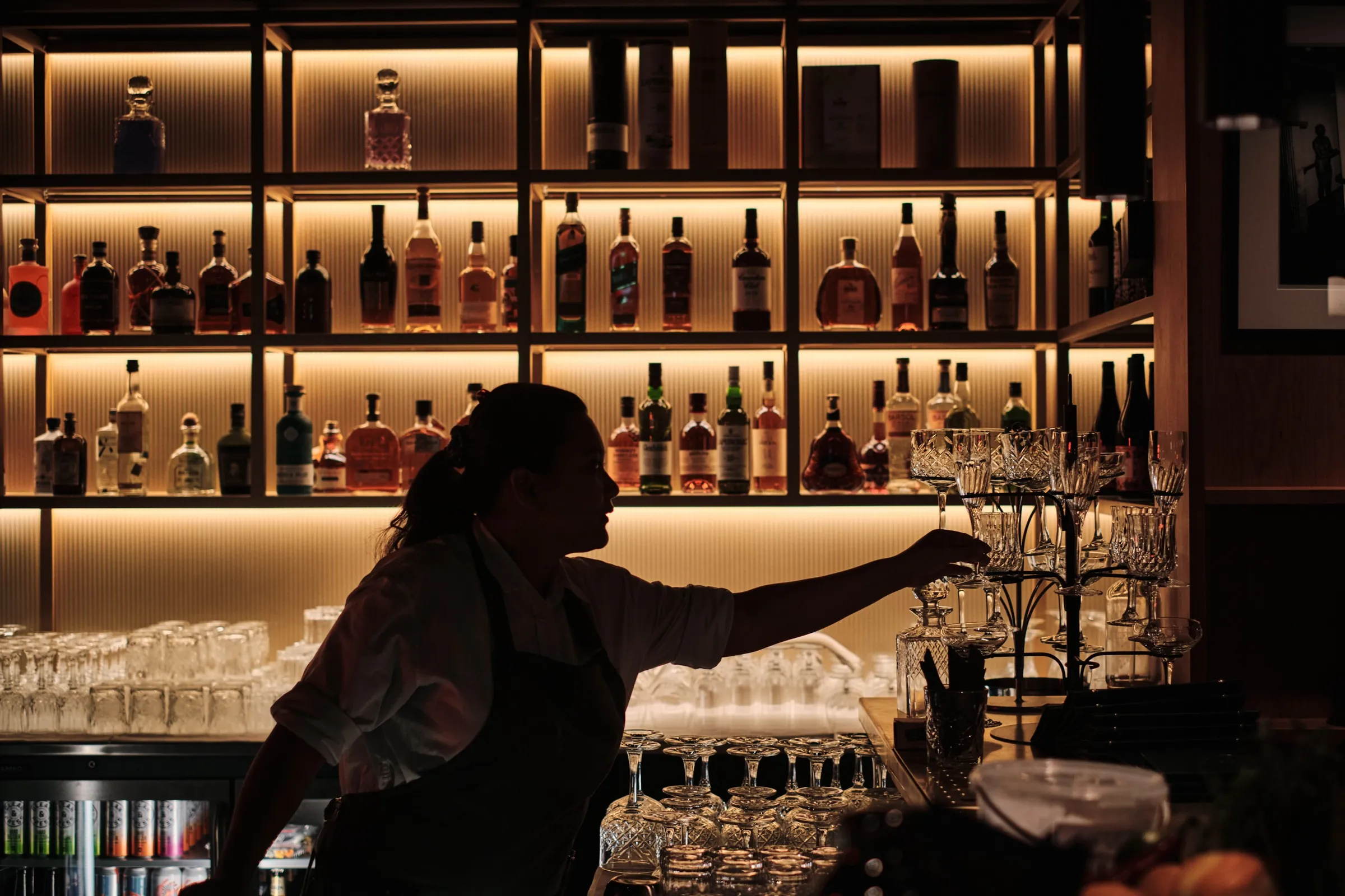 Bartender reaching for glasses in front of the back-lit bottle shelves at The Porter's Store