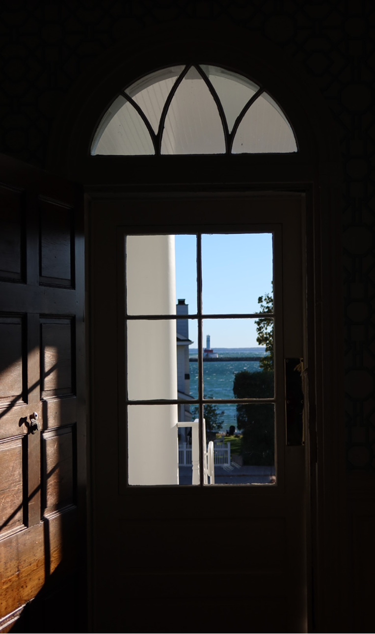 A dark wood doorway with an arched transom window, framing a bright view of white railings, trees and a lighthouse on the water outside.