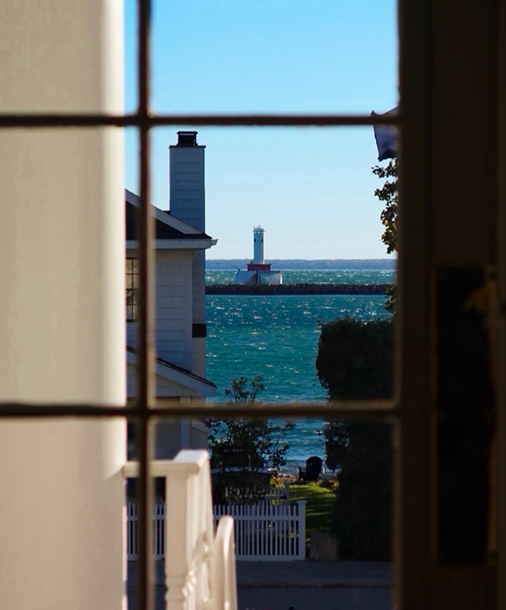 A view through a paned window past a white column and railing to a small lighthouse on sparkling blue water.