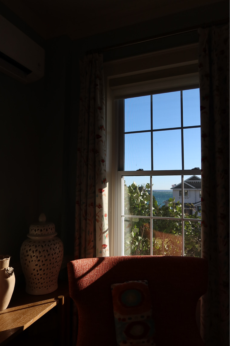 A dim room with floral curtains, a red armchair and a decorative ceramic jar beside a sunlit window looking out to trees, a house and the lake.
