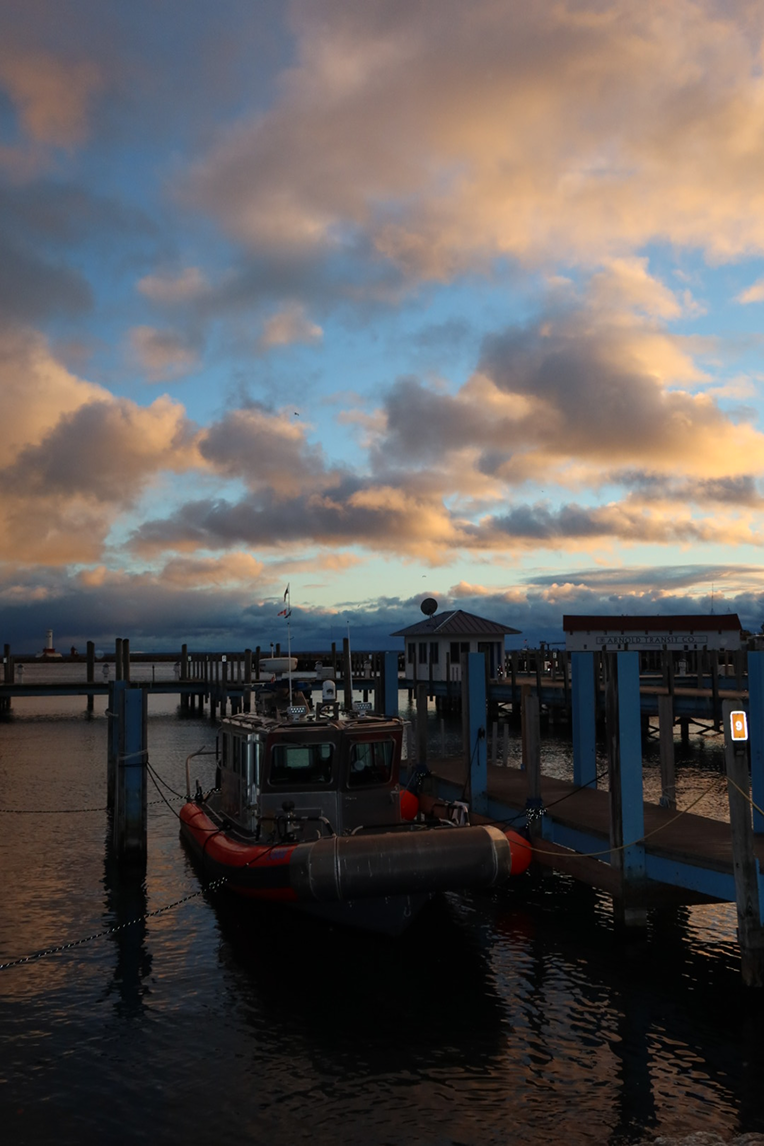A small patrol boat docked at a blue pier at dusk, with glowing orange clouds above the harbor.