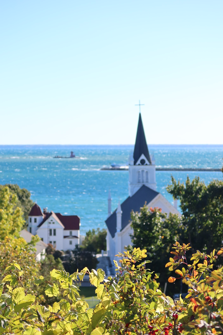 A white church with a dark steeple rising above green and orange foliage, with a bright blue lake and clear sky behind it.