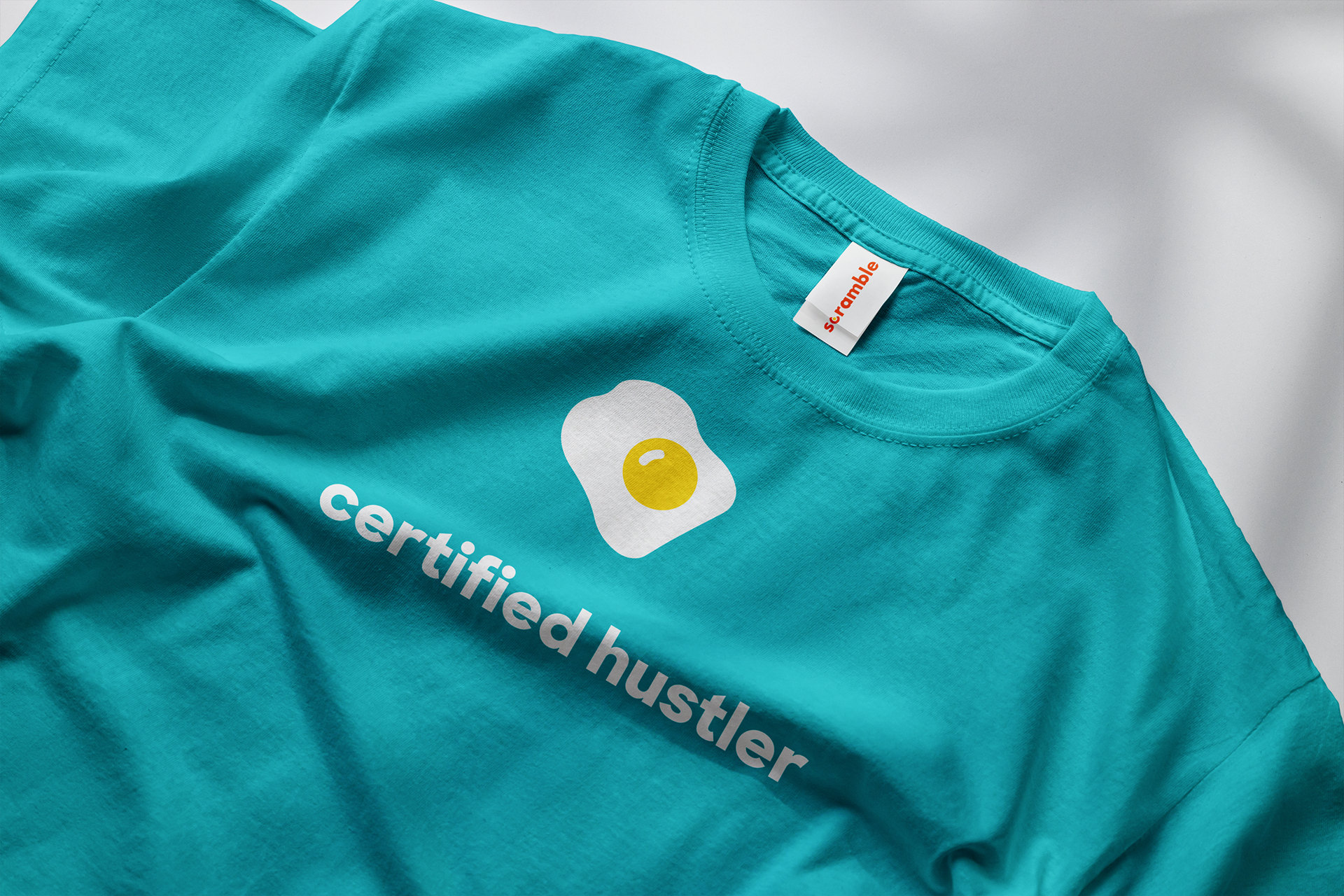 Teal Scramble t-shirt printed with a fried-egg mark and the words certified hustler, with a Scramble tag at the collar.