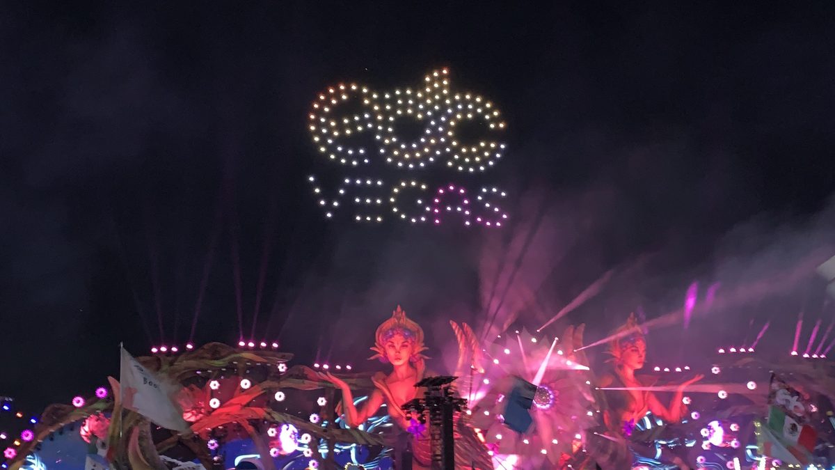 A drone show spells out 'EDC VEGAS' in glowing lights in the night sky above the kineticFIELD stage, with its giant illuminated figures and pink lasers below.