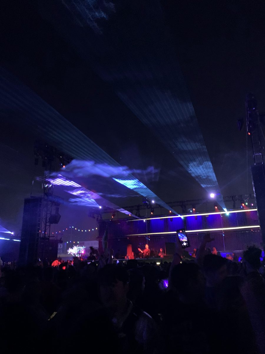 Blue laser beams fan out through haze above a smaller EDC Las Vegas stage at night, with a silhouetted crowd in the foreground.