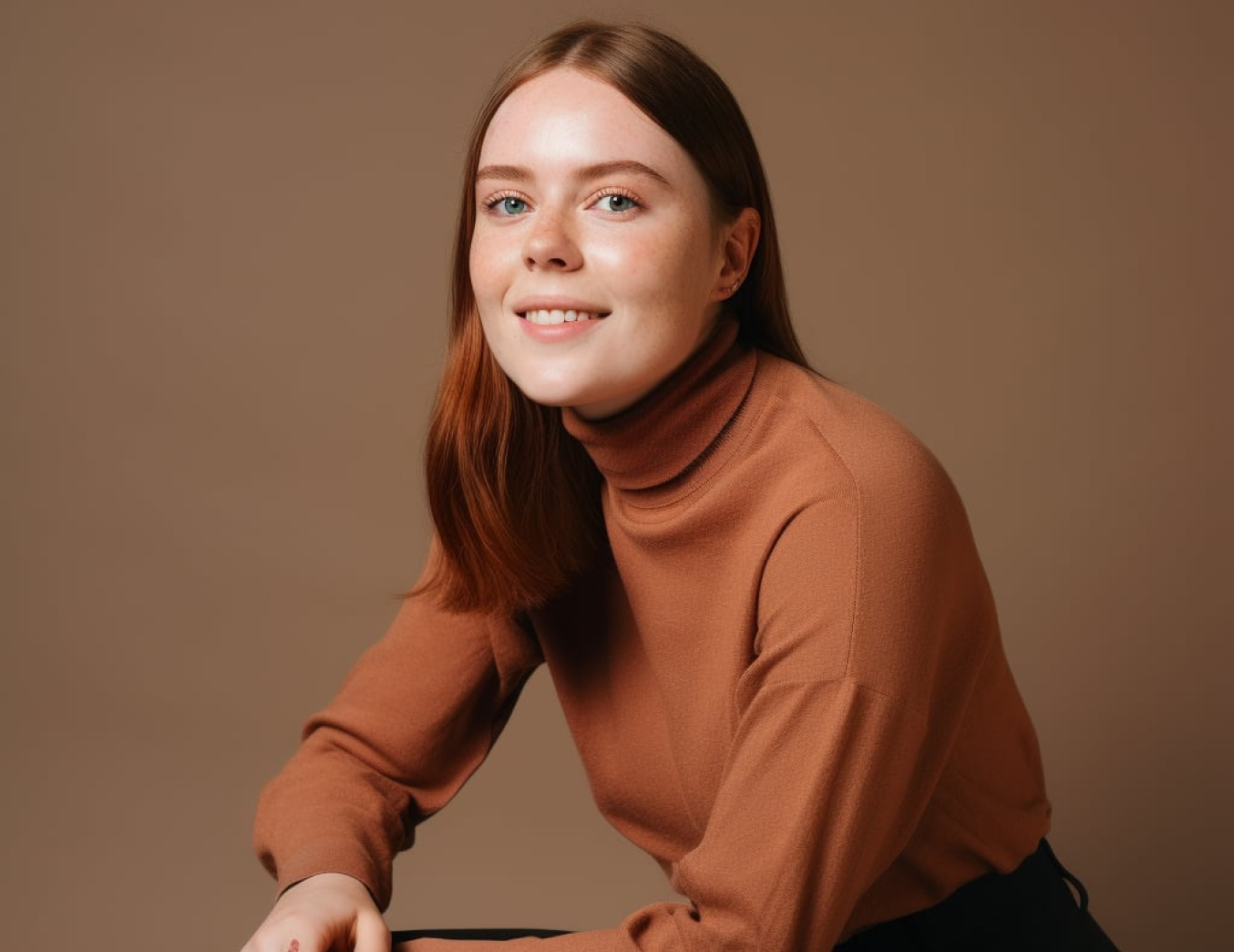 Young woman with long reddish-brown hair wearing a brown turtleneck sweater smiling against a brown background.