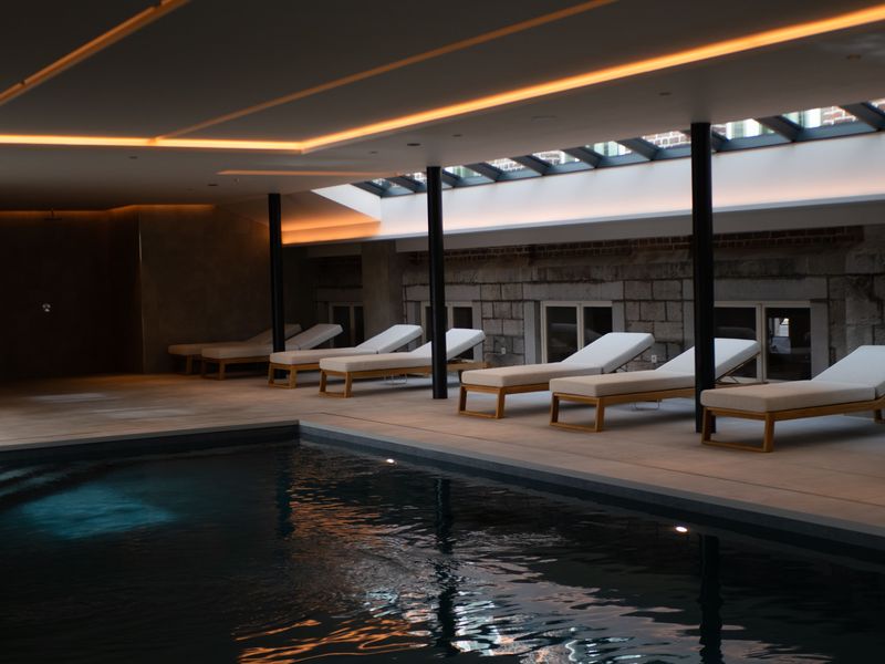 La piscine intérieure de DAO Verviers et ses transats blancs, sous des lignes de lumière chaude
