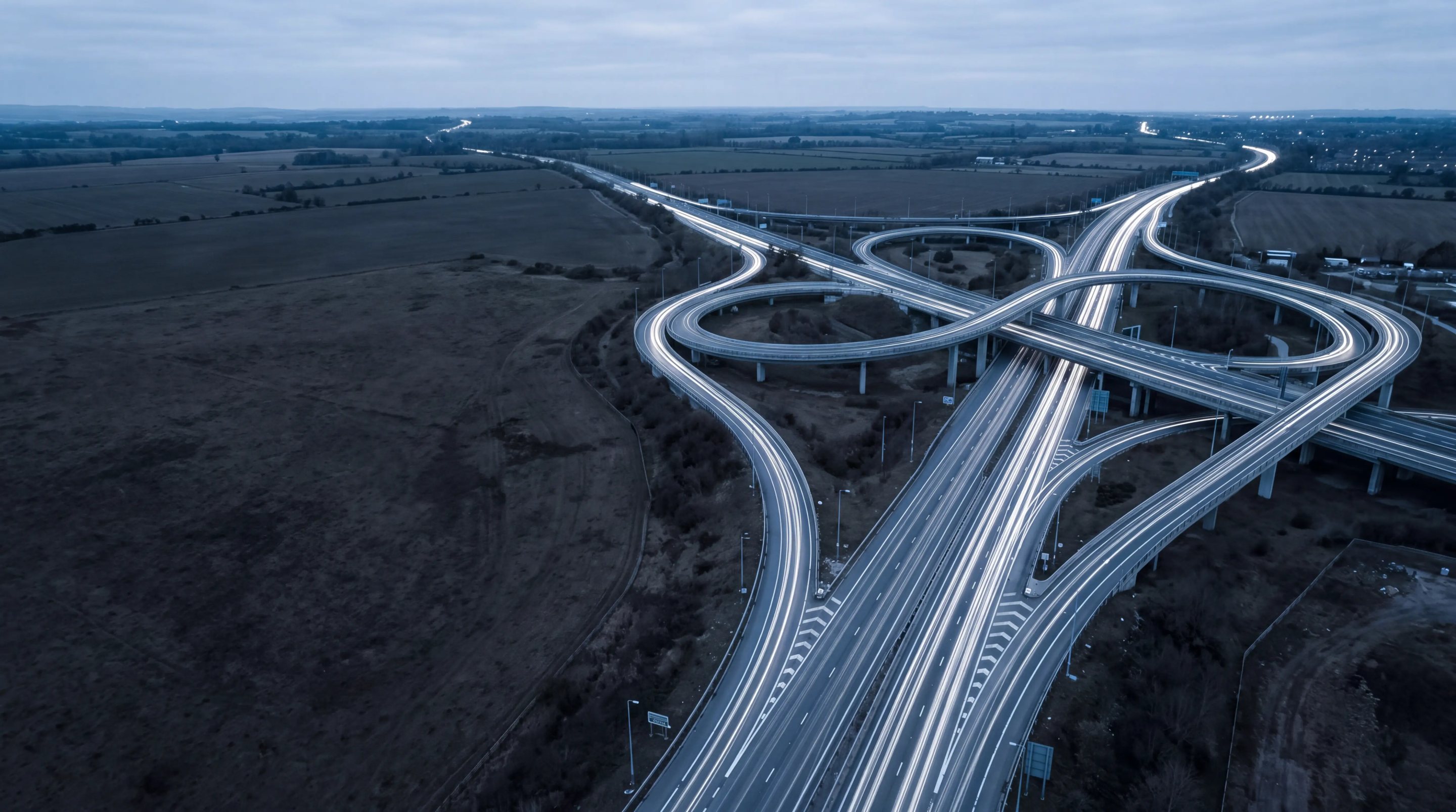A motorway interchange at dusk, traffic flowing through every ramp without stopping.