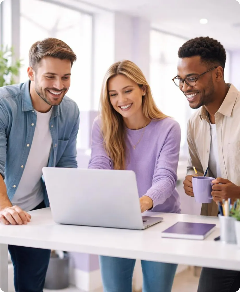 Three smiling coworkers gathered around a laptop at a desk in a bright office.