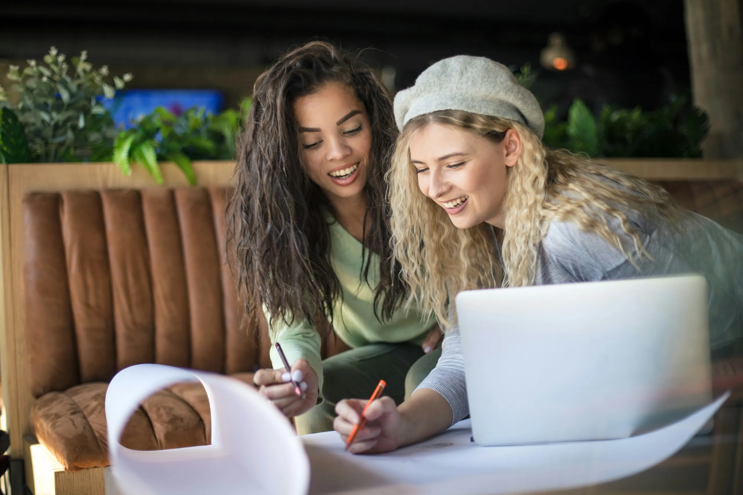 Two women lean over a large sheet of paper, smiling as they sketch out an idea together beside a laptop.