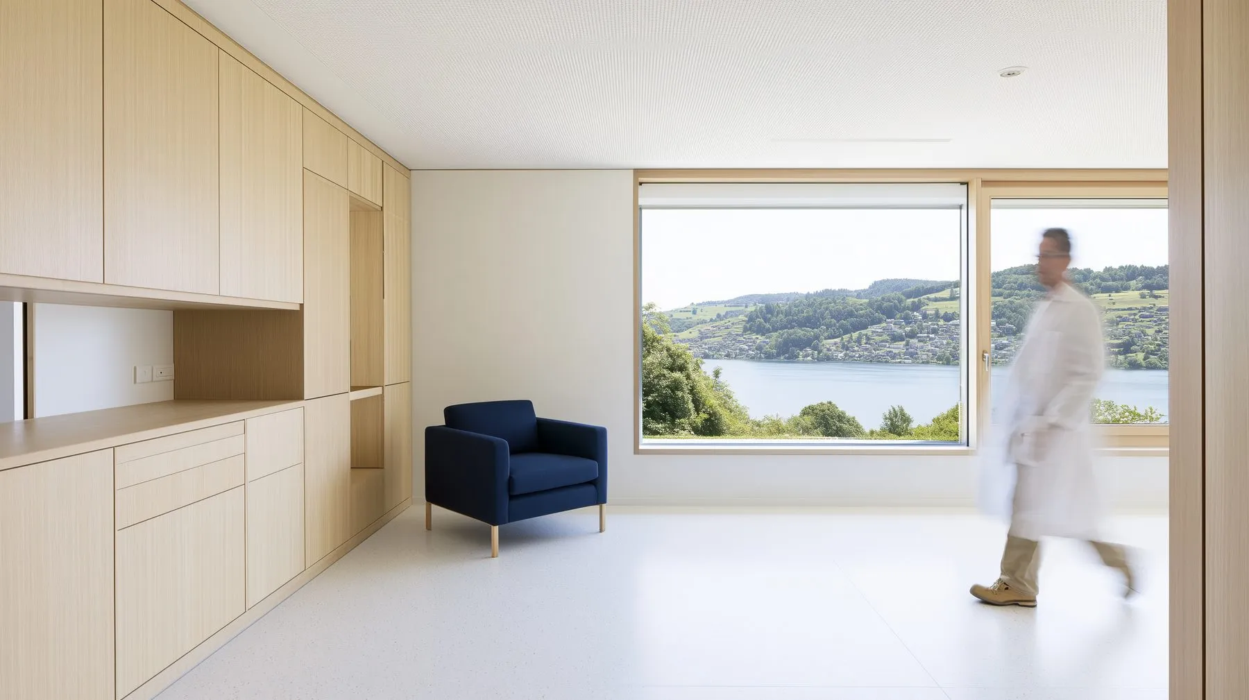 A calm consulting room with oak cabinets, a navy armchair and a large window onto a lake and green hills, a doctor walking through