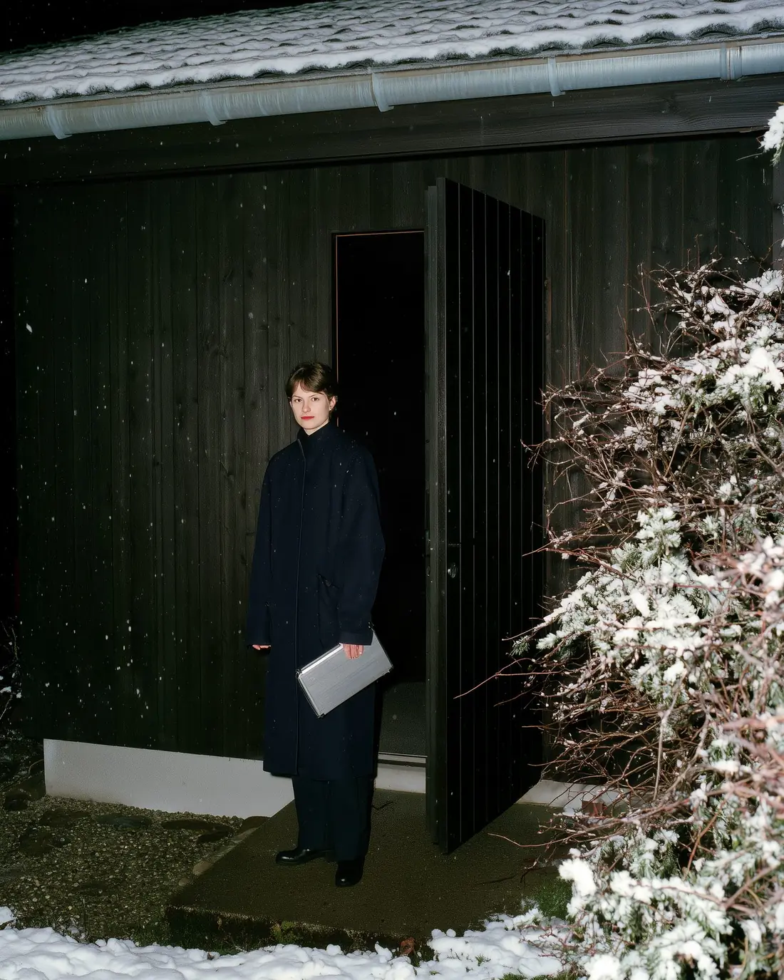 A nurse in a navy coat with an aluminium case at the open door of a timber house on a snowy night