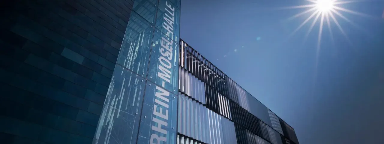 Von unten fotografierte Fassade der Rhein-Mosel-Halle in Koblenz mit senkrechter Beschriftung im Glas und Sonne über dem Dachrand